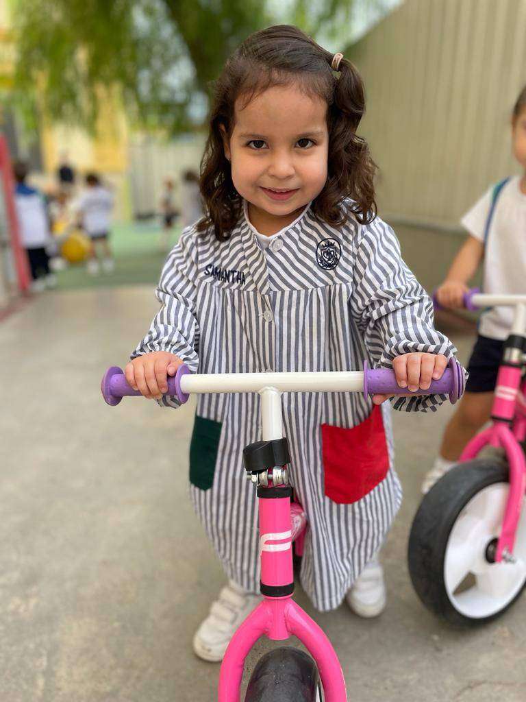 Niña morena con babero escolar Pureza de María Grao subida a una bicicleta rosa.